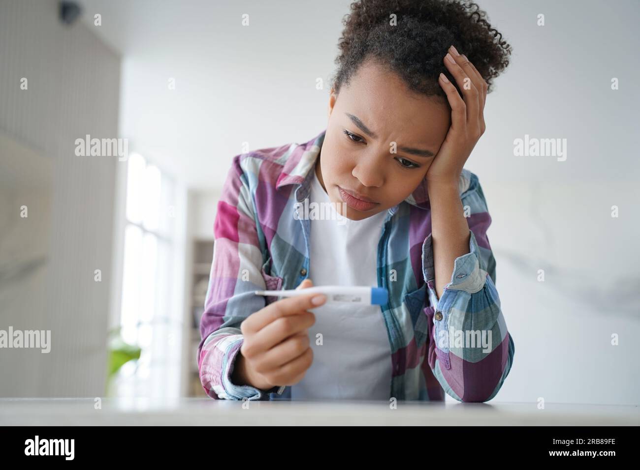 Une fille biraciale malsaine fronce les sourcils au thermomètre, déçue par la température élevée, affichant un symptôme de COVID. Banque D'Images