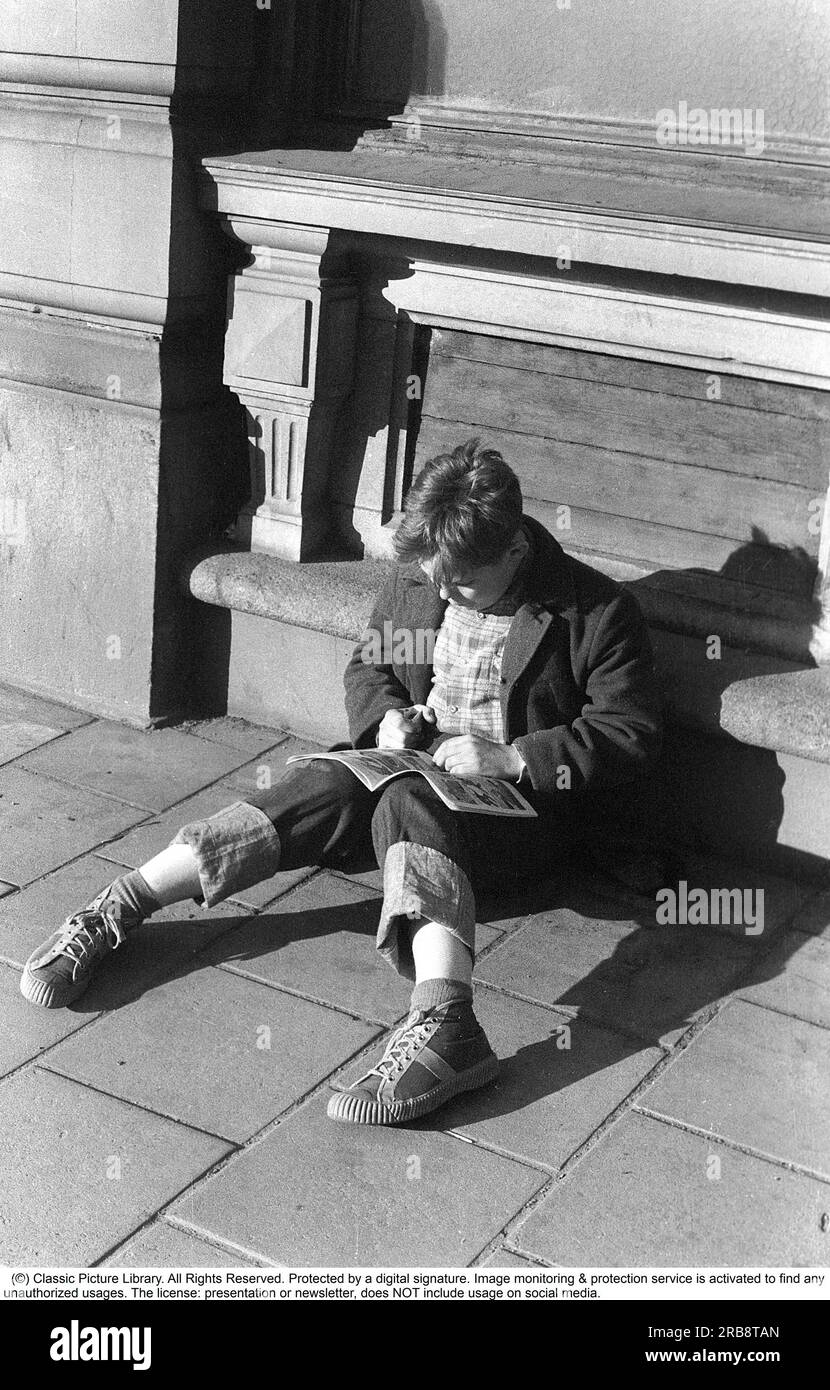 Stockholm dans les années 1950 Un garçon est assis à l'extérieur du bâtiment de l'éditeur Bonnier à Sveavägen 56 et lit une bande dessinée. L'année est 1953. Avec la sortie de Kalle Anka & C:o et Seriemagasinet en 1948, le phénomène de la bande dessinée a pris son envol en Suède. Après un boom dans les années 1950, le développement a quelque peu ralenti, en partie parce que le débat sur les séries américaines a également atteint la Suède. Au cours des années 1950 et 60, les bandes dessinées ont commencé à être considérées comme de la « littérature inappropriée » et ont été accusées de promouvoir la violence chez les enfants et les adolescents. Au lieu de cela, la lecture de livres a été saluée dans le général social d Banque D'Images