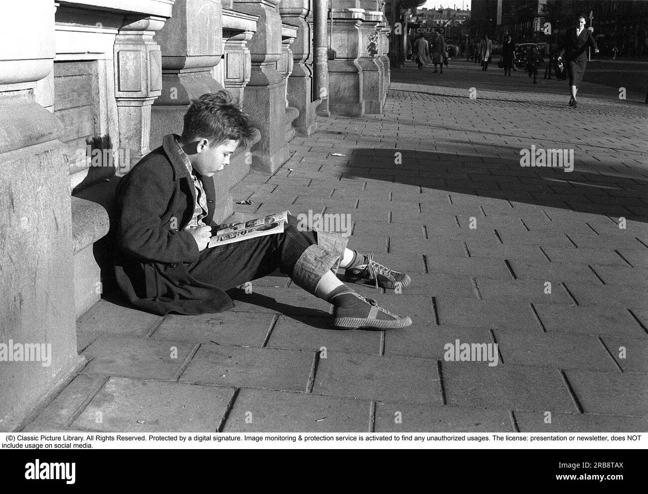 Stockholm dans les années 1950 Un garçon est assis à l'extérieur du bâtiment de l'éditeur Bonnier à Sveavägen 56 et lit une bande dessinée. L'année est 1953. Avec la sortie de Kalle Anka & C:o et Seriemagasinet en 1948, le phénomène de la bande dessinée a pris son envol en Suède. Après un boom dans les années 1950, le développement a quelque peu ralenti, en partie parce que le débat sur les séries américaines a également atteint la Suède. Au cours des années 1950 et 60, les bandes dessinées ont commencé à être considérées comme de la « littérature inappropriée » et ont été accusées de promouvoir la violence chez les enfants et les adolescents. Au lieu de cela, la lecture de livres a été saluée dans le général social d Banque D'Images