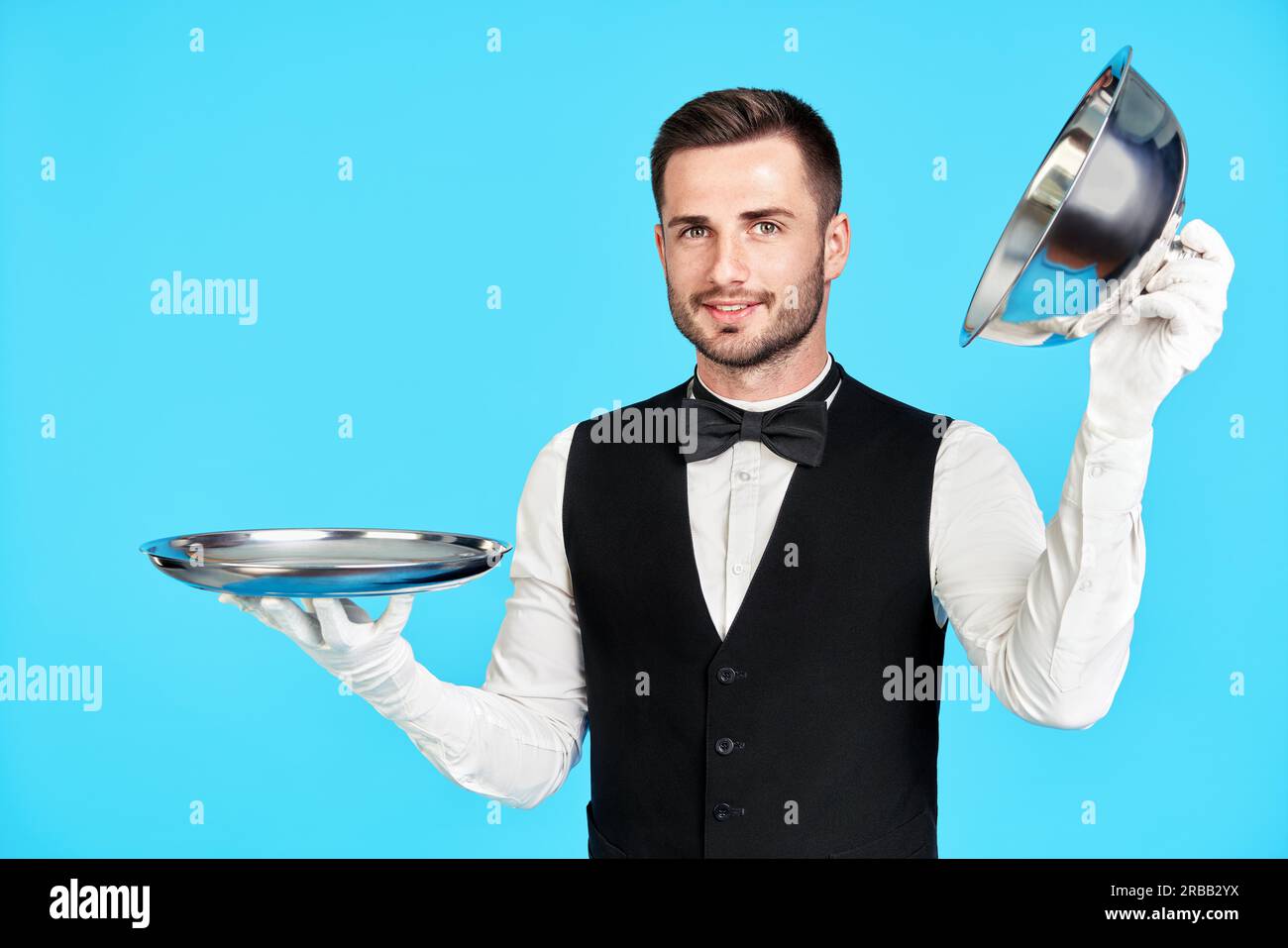 Élégant jeune serveur tenant le cloche sur un plateau vide prêt à servir sur fond bleu. Restaurant, horeca, concept de service de première classe Banque D'Images