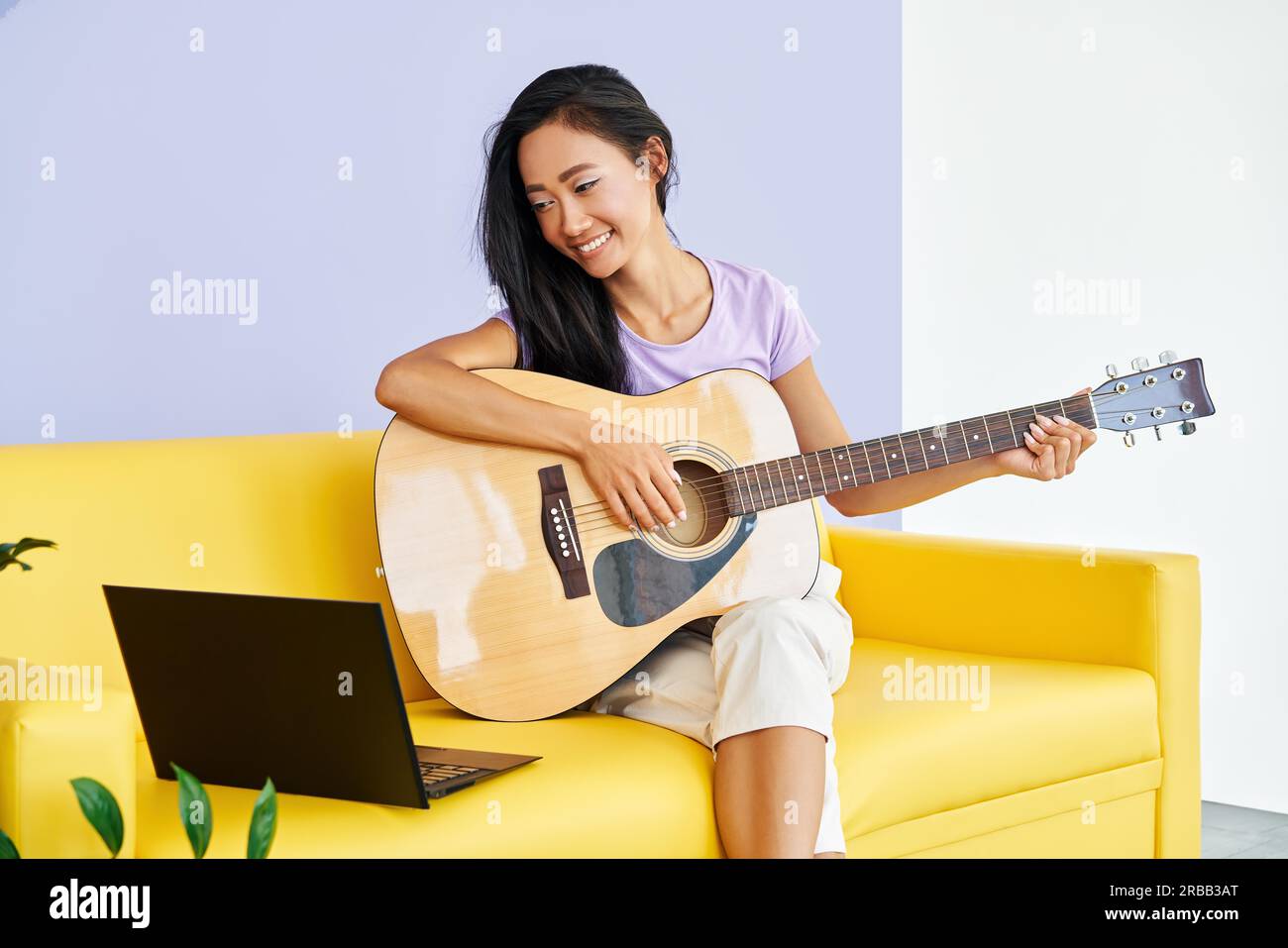 Sourire jolie femme apprenant à jouer de la guitare regarde une leçon vidéo en ligne à l'aide d'un ordinateur portable. Formation en ligne, hobby, concept musical Banque D'Images