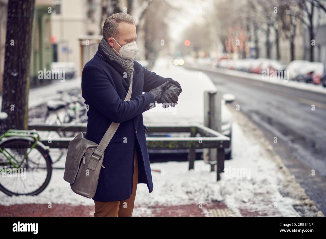 Homme d’affaires avec sac à bandoulière portant un masque facial pendant la pandémie de Covid-19 debout dans une rue urbaine enneigée en hiver vérifiant l’heure sur le sien Banque D'Images