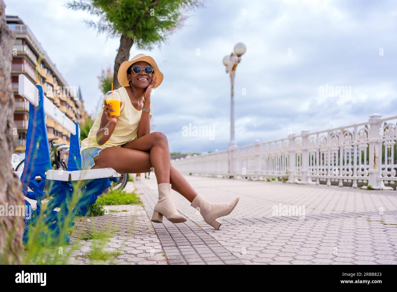 Femme ethnique noire ayant un jus assis sur un banc dans la ville, touriste profitant de l'été Banque D'Images