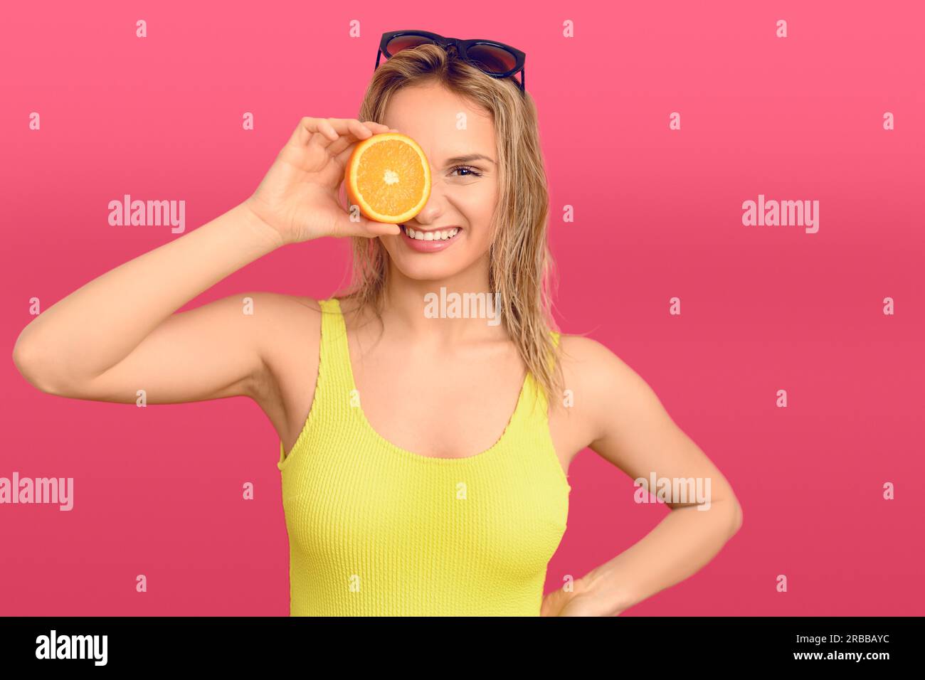 Heureuse jeune femme en bonne santé tenant un orange frais de moitié juteux à ses yeux avec un sourire amical et beaming sur un fond de studio rose Banque D'Images