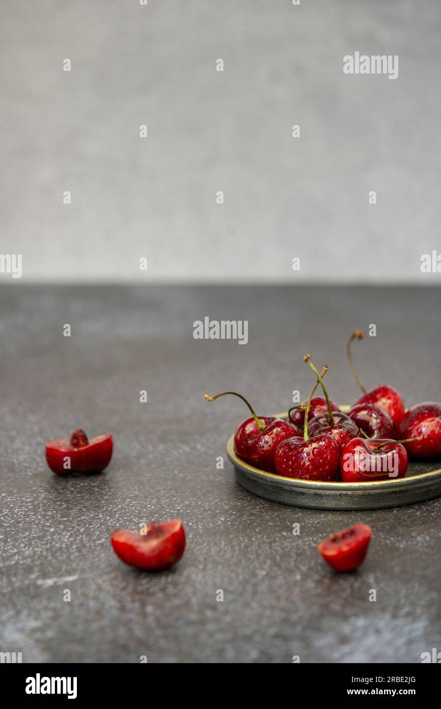 Quelques délicieuses cerises mûres dans une petite soucoupe en métal bordée d'or, certaines coupées sur une surface sombre Banque D'Images