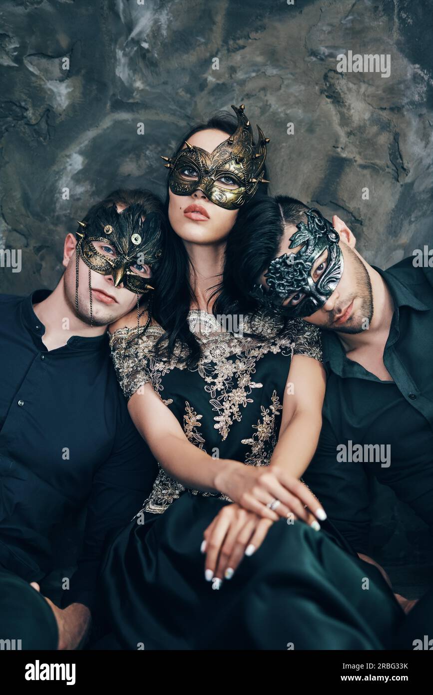 Belle jeune femme et deux hommes de mystérieux masque noir assis sur le plancher en studio. La mode, concept parti Banque D'Images