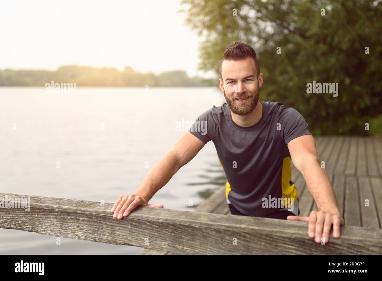 Jeune homme de faire des exercices d'étirement sur une terrasse en bois donnant sur un lac comme il sourit à la caméra dans un concept de vie sain Banque D'Images