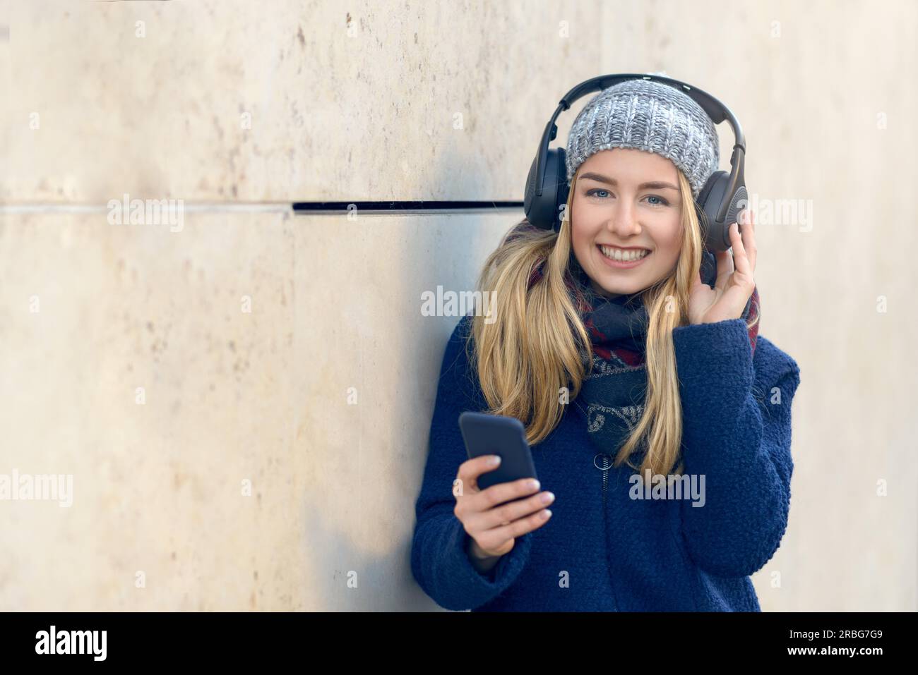 Jeune femme blonde en manteau et chapeau debout à l'extérieur contre le mur de béton tenant le téléphone intelligent en main avec les yeux fermés souriant écouter de la musique dans Banque D'Images