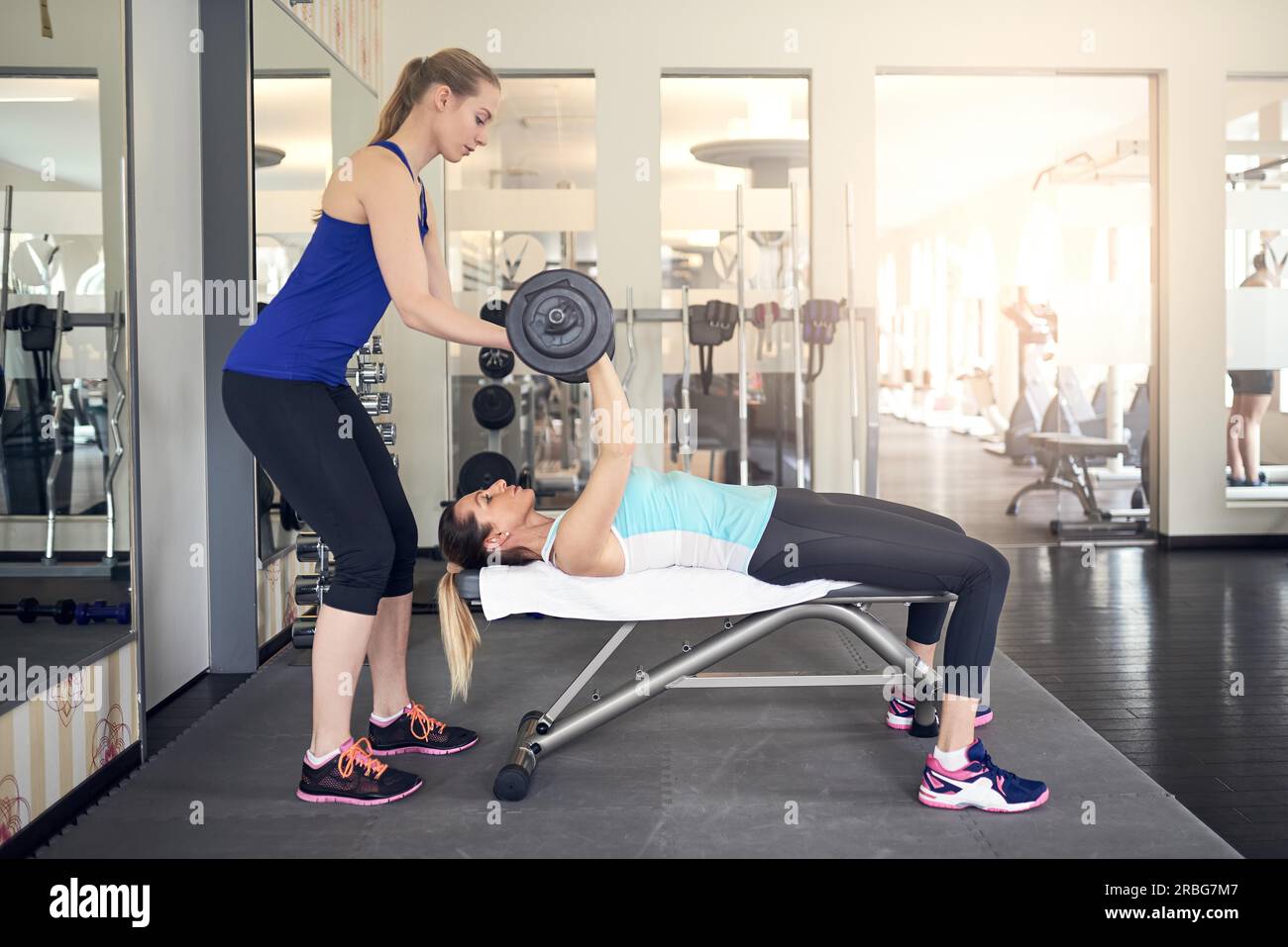 Jeune femme travaillant avec des poids d'haltères sur un banc dans la salle de sport avec l'aide de son entraîneur personnel dans un concept de santé et de remise en forme Banque D'Images