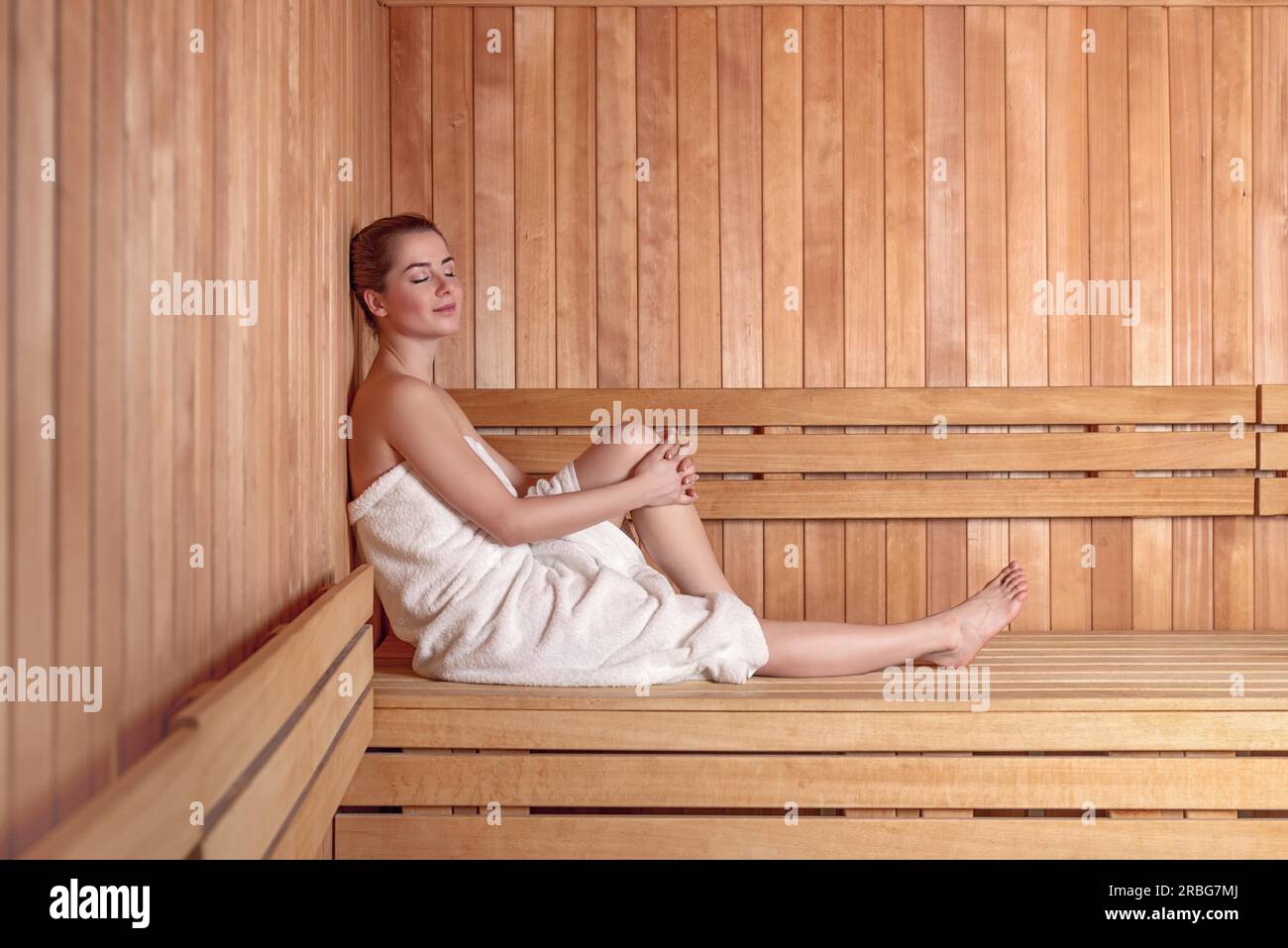 Side view full length portrait of young woman smiling sitting on bench in sauna en bois canapé enveloppé dans une serviette blanche, avec des cheveux en chignon donut Banque D'Images