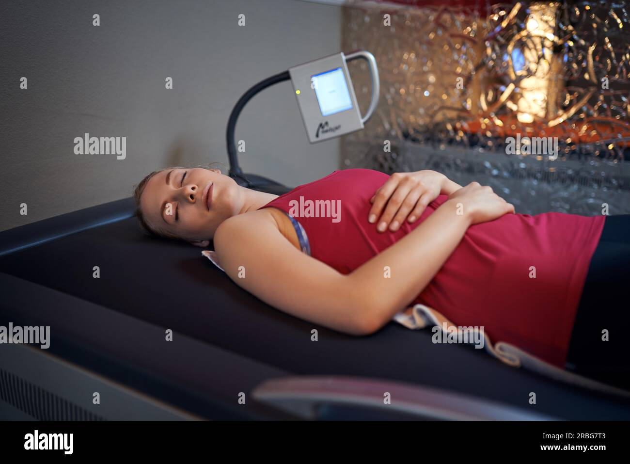Woman in red shirt portant sur son dos sur table en salle de massage avec écran à côté de sa tête et de verre mur derrière, looking at camera Banque D'Images