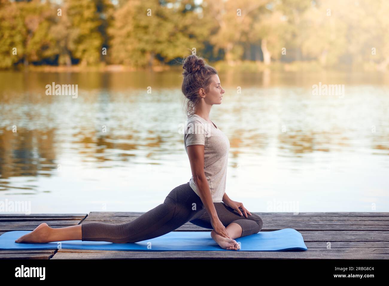 Attractive young woman working out on mat sur une terrasse en bois au-dessus d'un lac ou rivière sitting avec ses yeux fermés et une expression sereine Banque D'Images