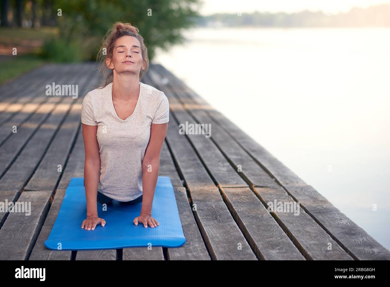 Attractive young woman working out on mat sur une terrasse en bois au-dessus d'un lac ou rivière sitting avec ses yeux fermés et une expression sereine Banque D'Images