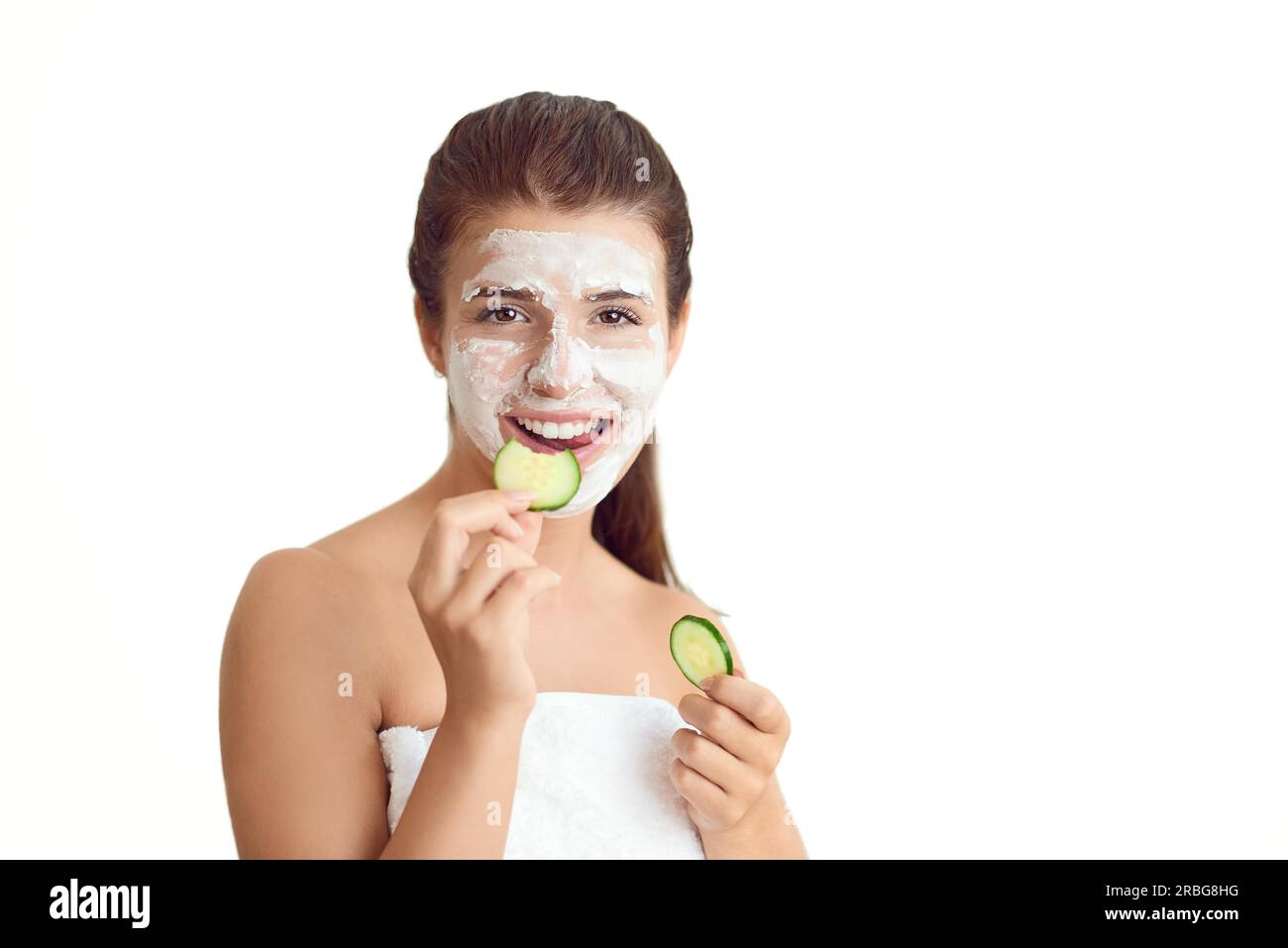 Souriant jeune femme attrayante dans une serviette portant un traitement de masque facial tenant des tranches de concombre cru pour ses yeux dans un concept de spa et de beauté Banque D'Images