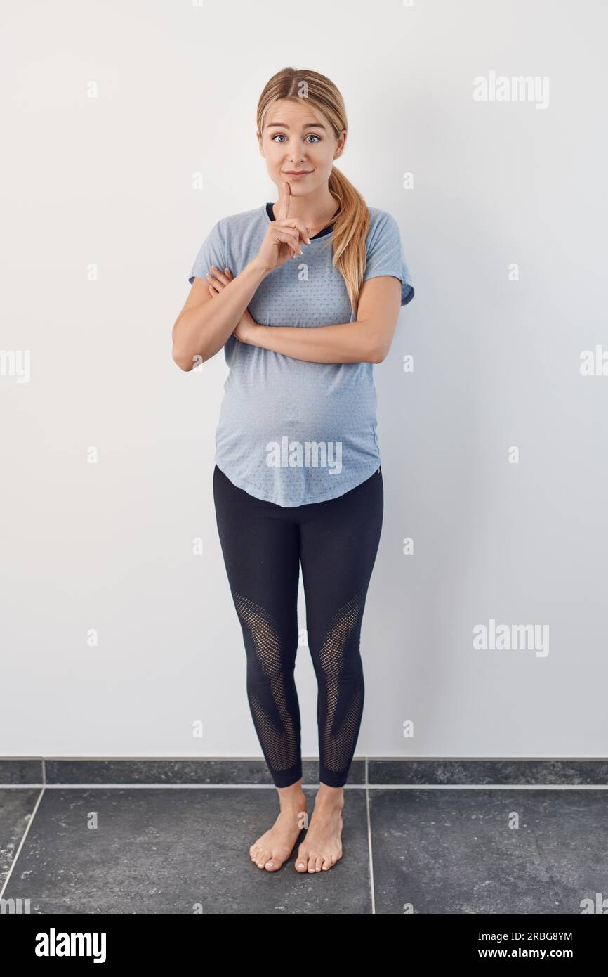 Surprise pieds nus jeune femme enceinte debout avec sa main à son menton levant ses sourcils avec de larges yeux debout sur toute la longueur contre un blanc Banque D'Images