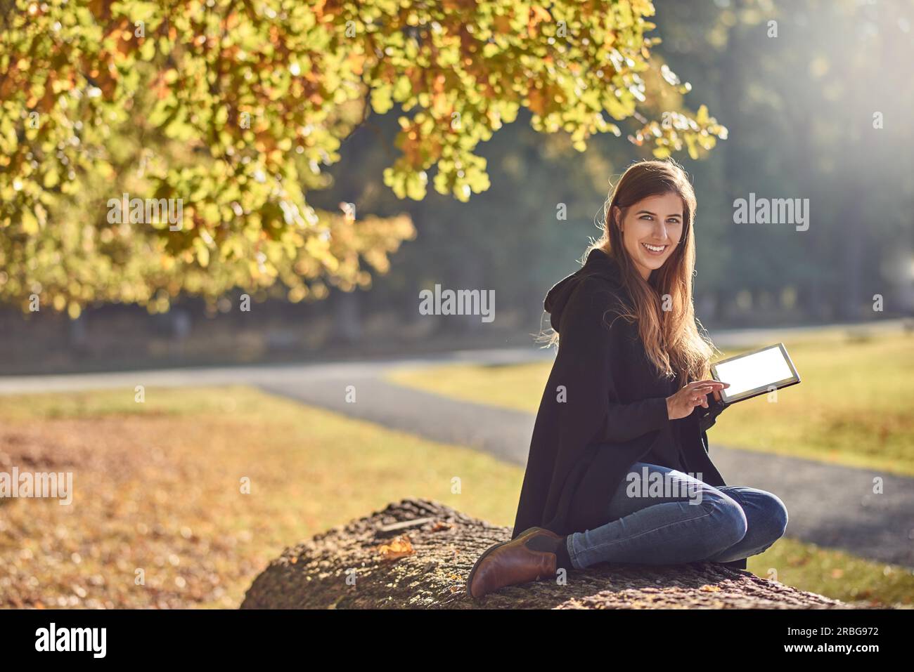 Pretty young woman relaxing in an autumn park lecture sur son PC tablette et sourit joyeusement à l'appareil photo Banque D'Images