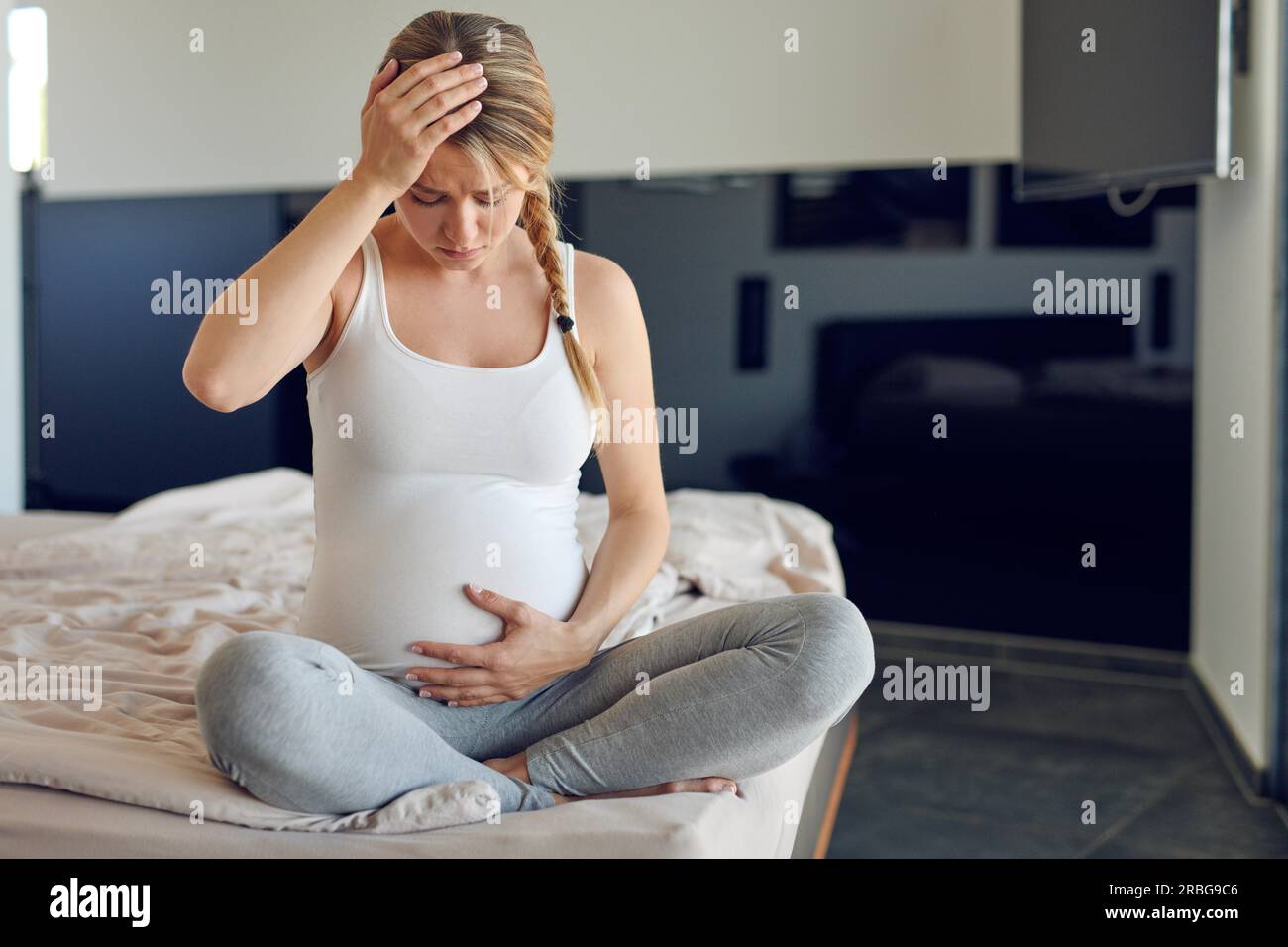 Trouble très enceinte jeune femme assise jambes croisées sur un lit berçant son abdomen enflé et serrant sa tête regardant vers le bas avec un sérieux Banque D'Images