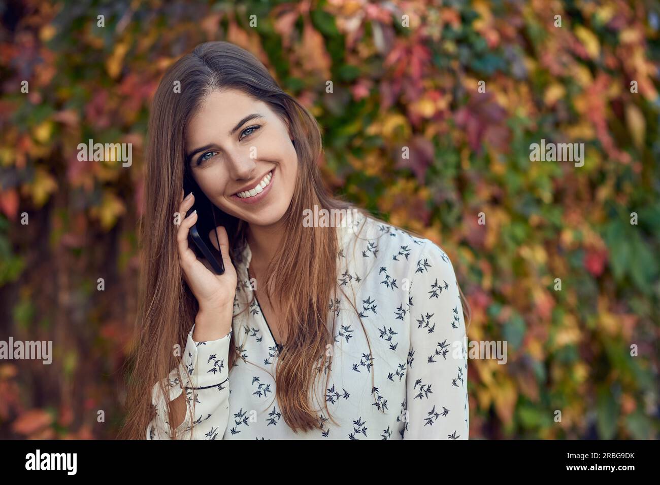 Souriante jeune femme chat sur un mobile à l'automne, debout devant un réducteur coloré souriant joyeusement à l'appareil photo avec sun flare Banque D'Images