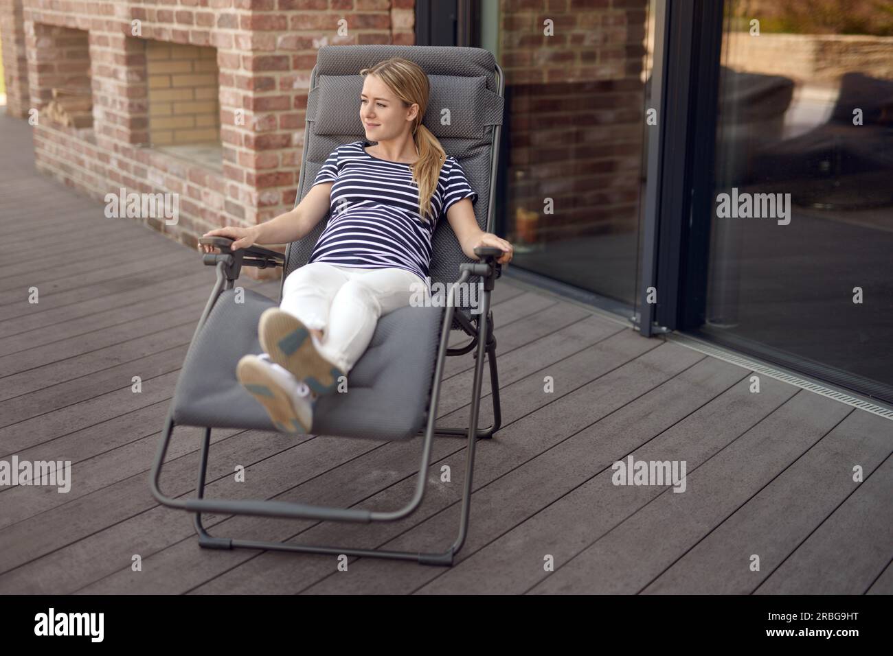 Happy smiling attractive young pregnant woman détente en plein air sur un fauteuil inclinable sur une terrasse en bois en berçant son bébé bosse dans ses mains Banque D'Images