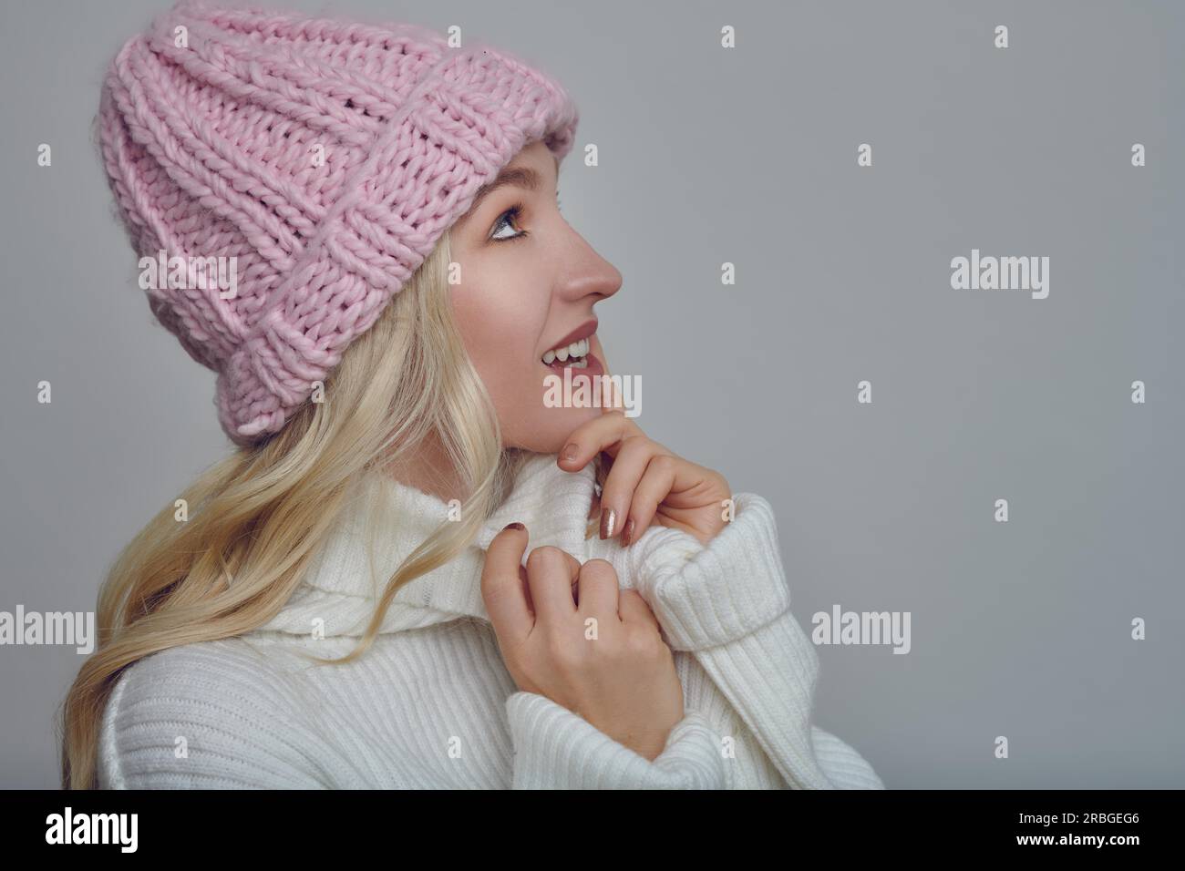 Jolie jeune femme blonde en tenue d'hiver lainée câlinant dans la chaleur de son pull col polo avec un sourire heureux tout en portant un tricot rose Banque D'Images