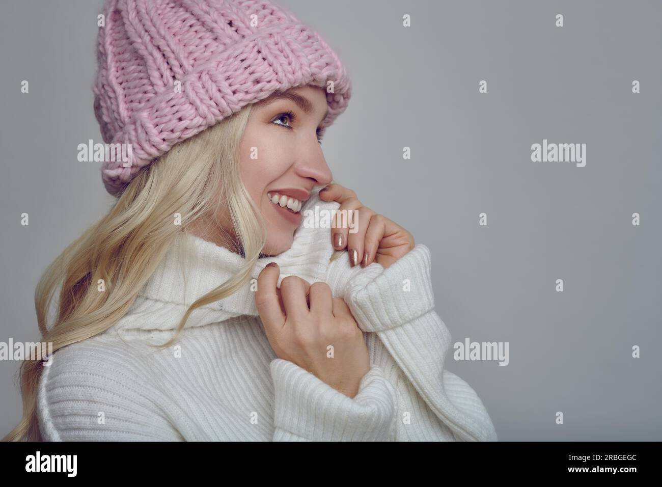 Jolie jeune femme blonde en tenue d'hiver lainée câlinant dans la chaleur de son pull col polo avec un sourire heureux tout en portant un tricot rose Banque D'Images