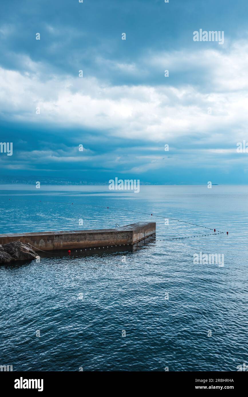 Jetée en pierre de béton à la ville de Lovran dans le golfe de Kvarner de la mer Adriatique au coucher du soleil avec ciel couvert de tempête Banque D'Images