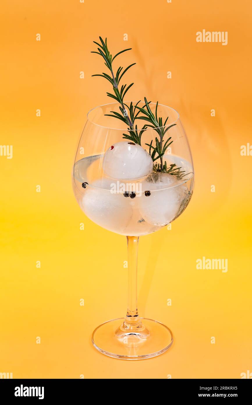 Photo studio d'un cocktail gin tonic avec romarin, poivre et glaçons isolé en verre sur fond jaune frais Banque D'Images