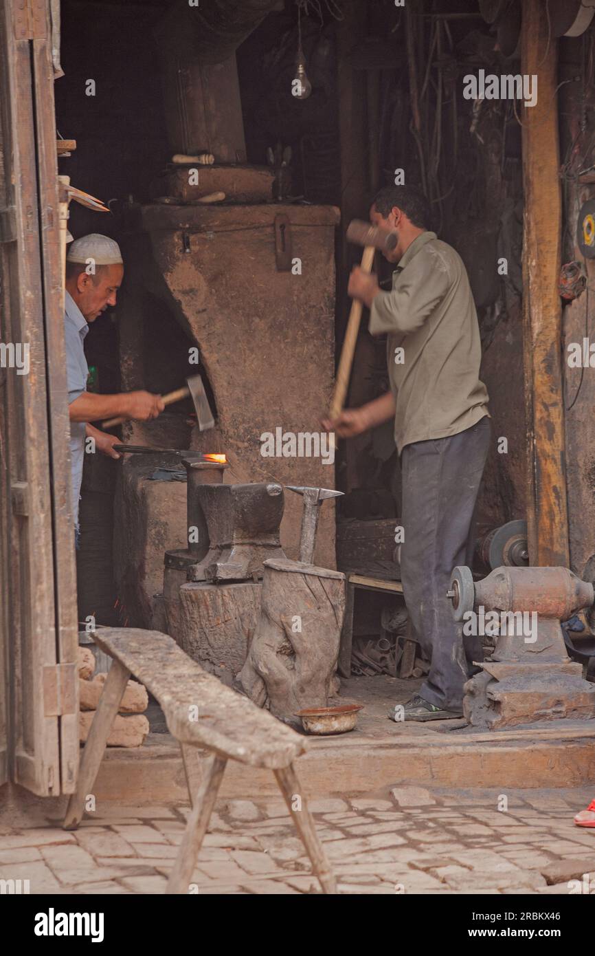 Deux forgerons travaillant dans un atelier en bord de route sur les marchés de Kashgar. L'un travaillant au four et l'autre utilisant un marteau de forgeron Banque D'Images