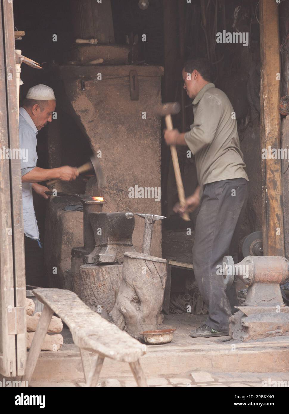 Deux forgerons travaillant dans un atelier en bord de route sur les marchés de Kashgar. L'un travaillant au four et l'autre utilisant un marteau de forgeron Banque D'Images