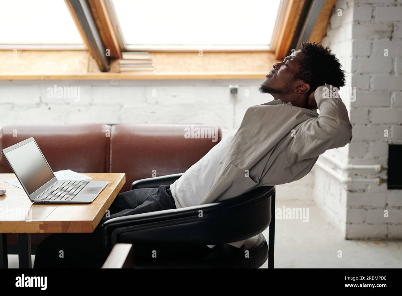 Jeune homme d'affaires afro-américain surmené assis dans un fauteuil en cuir devant un ordinateur portable au bureau et étirant les muscles du corps Banque D'Images
