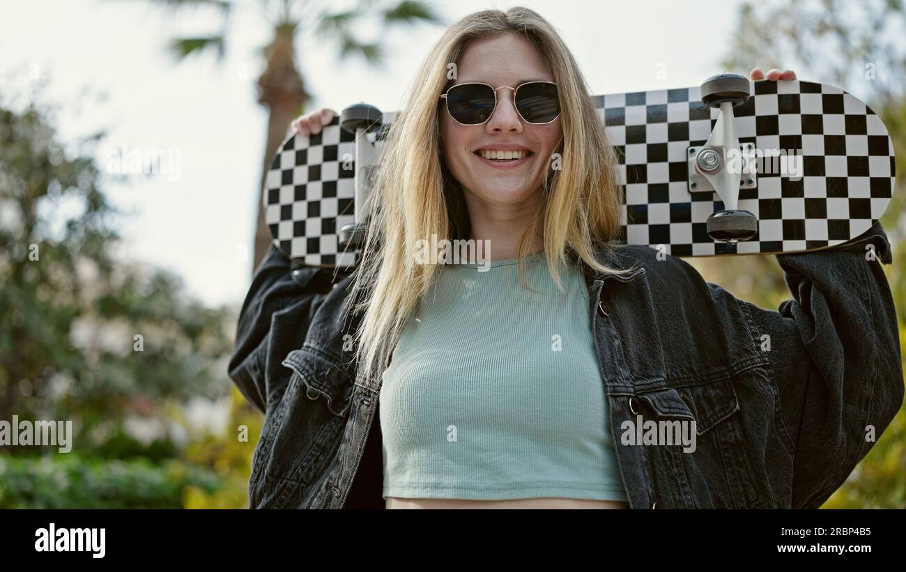 Jeune femme blonde souriante confiante tenant le skate au parc Banque D'Images