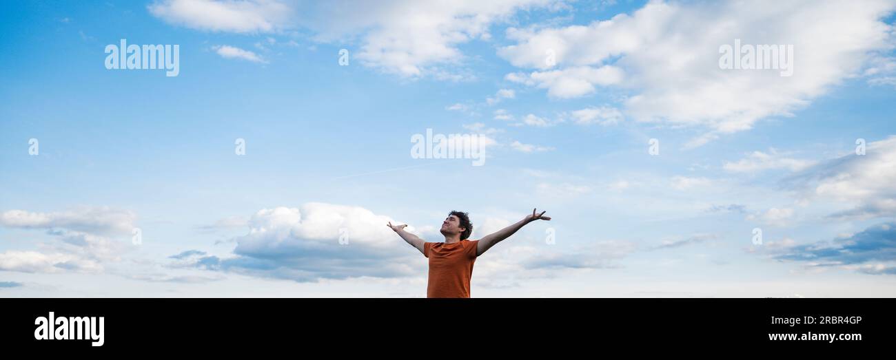 Image vue large d'un jeune homme pratiquant la pleine conscience ou méditant debout avec ses bras étendus sous le ciel bleu avec des nuages blancs. Banque D'Images