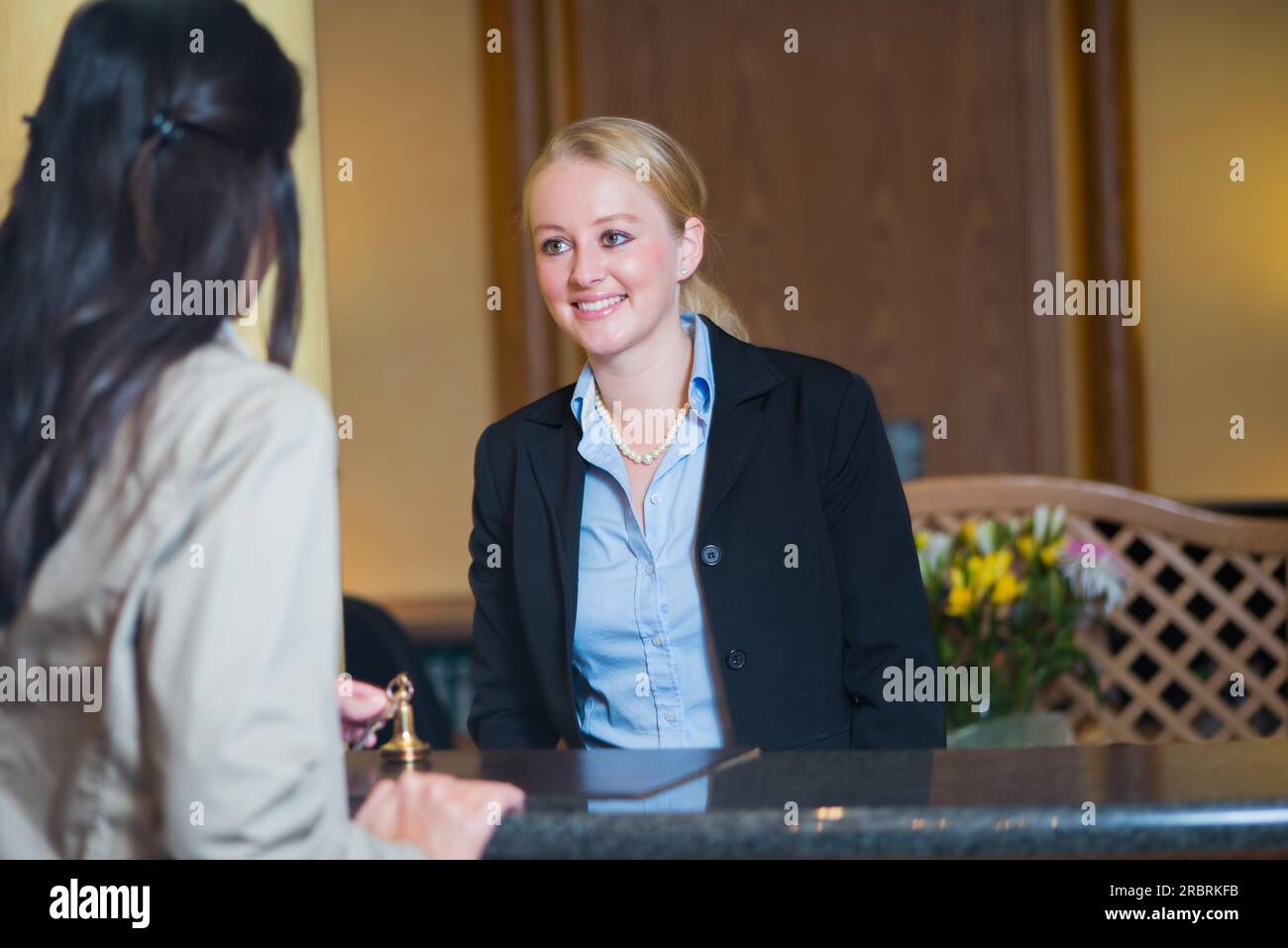 Belle réceptionniste blonde élégante se tenant derrière le comptoir de service dans un hall d'hôtel regardant un client de l'hôtel avec un sourire amical Banque D'Images