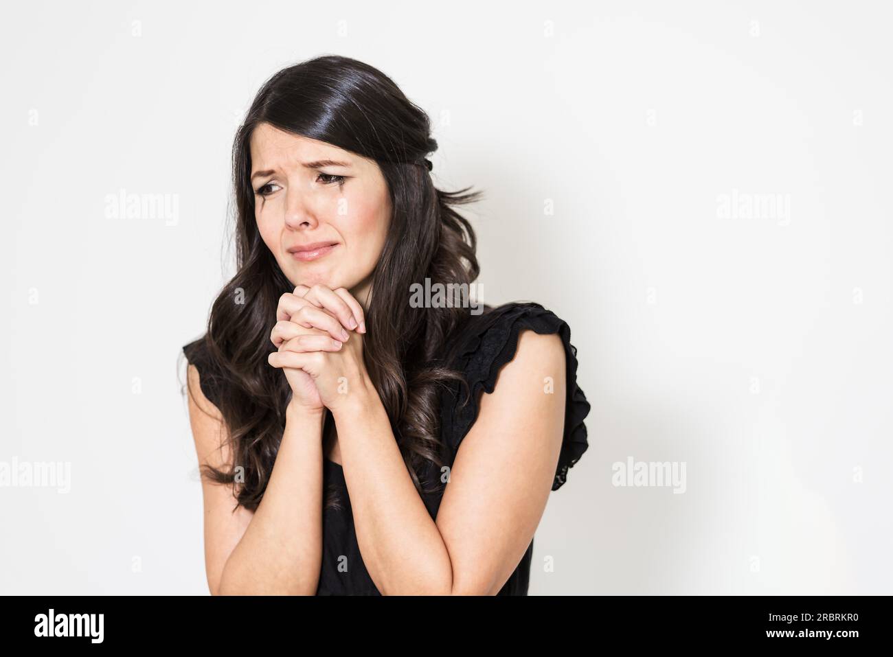 Jeune femme en pleurs éperdus avec elle les mains jointes dans l'angoisse debout avec les yeux baissés contre un fond studio blanc Banque D'Images