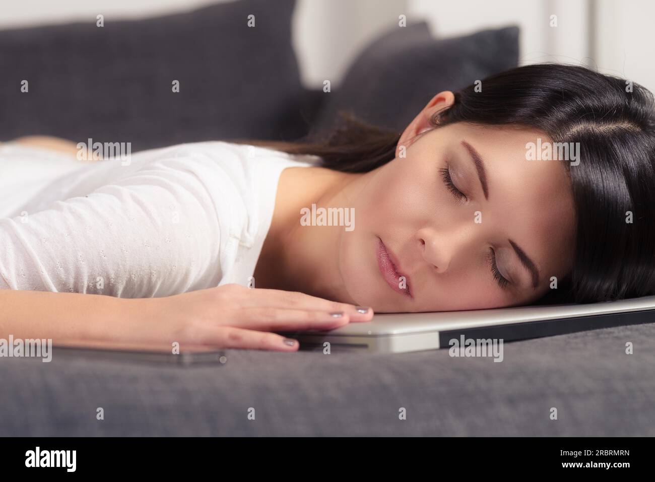 Jolie jeune femme posant sa tête sur un ordinateur portable avec ses yeux fermés, profitant d'un sommeil paisible sur le canapé Banque D'Images