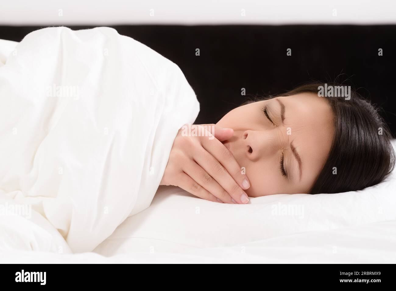 Fatiguée jeune femme attrayante couchée dans le lit bâillant avec sa main à sa bouche et les yeux fermés alors qu'elle se prépare à aller dans un sommeil rafraîchissant pour se détendre Banque D'Images