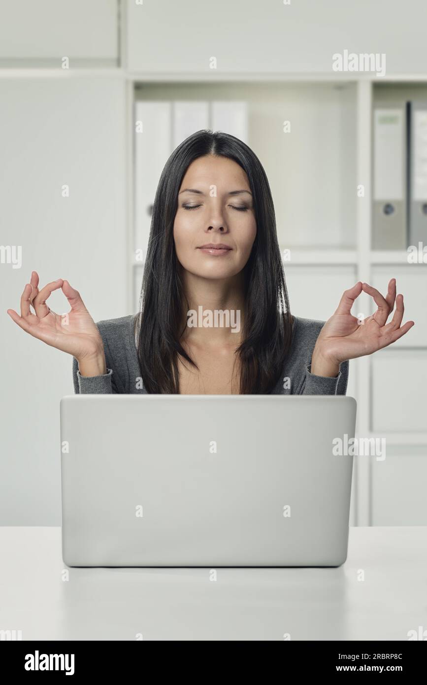 Close up Pretty Young Woman à l'aide d'un ordinateur portable pensant avec les deux mains dans les gestes de yoga et les yeux sont fermés, dans le bureau Banque D'Images