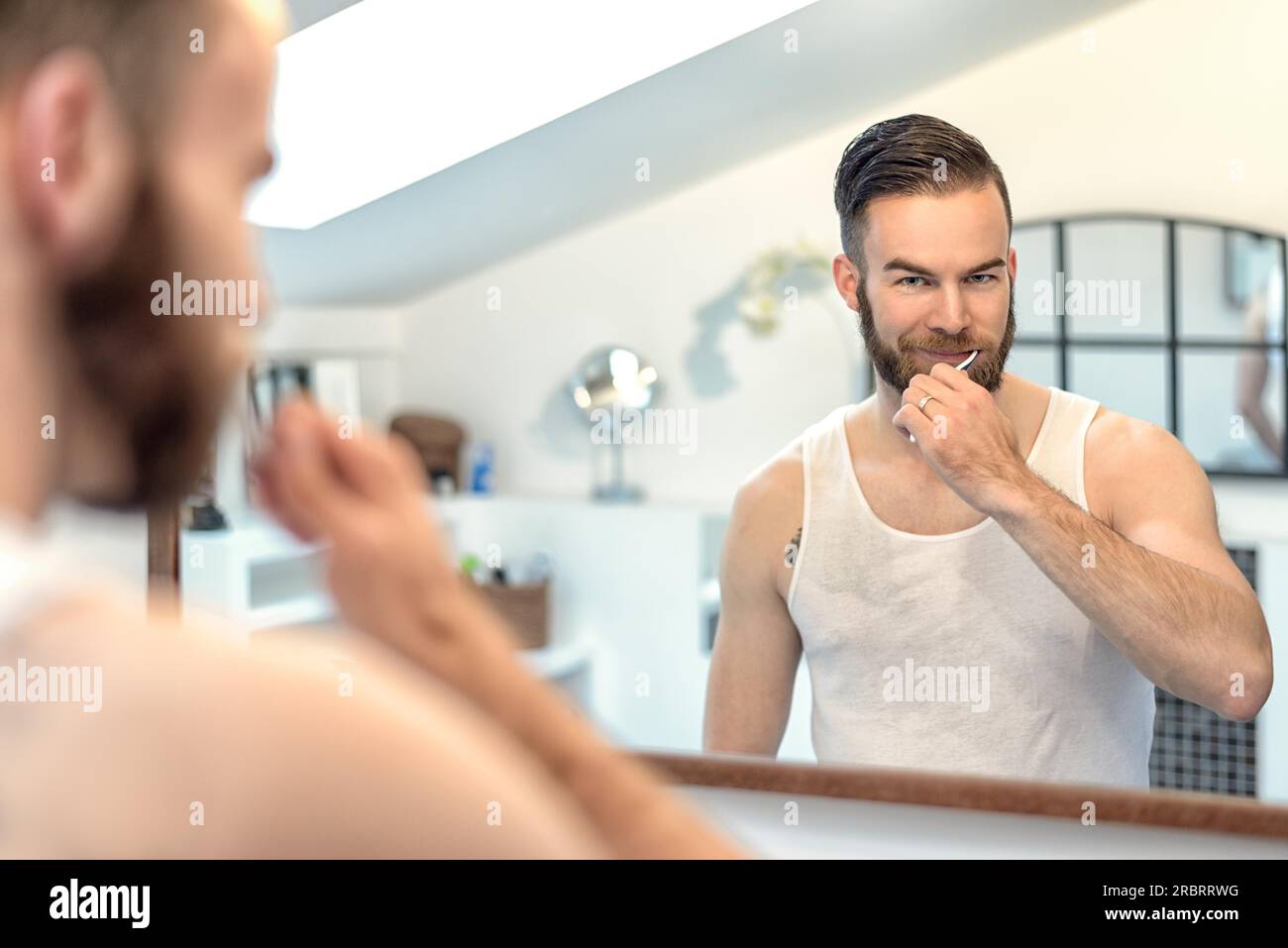 Séduisant jeune homme barbu se brosser les dents dans la salle de bains à regarder dans le miroir dans un concept d'hygiène dentaire Banque D'Images