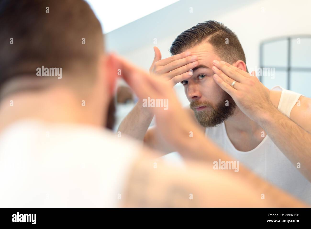 Homme barbu vérifiant son teint dans le miroir de la salle de bain, regardant inquiet, vue arrière sur l'épaule Banque D'Images