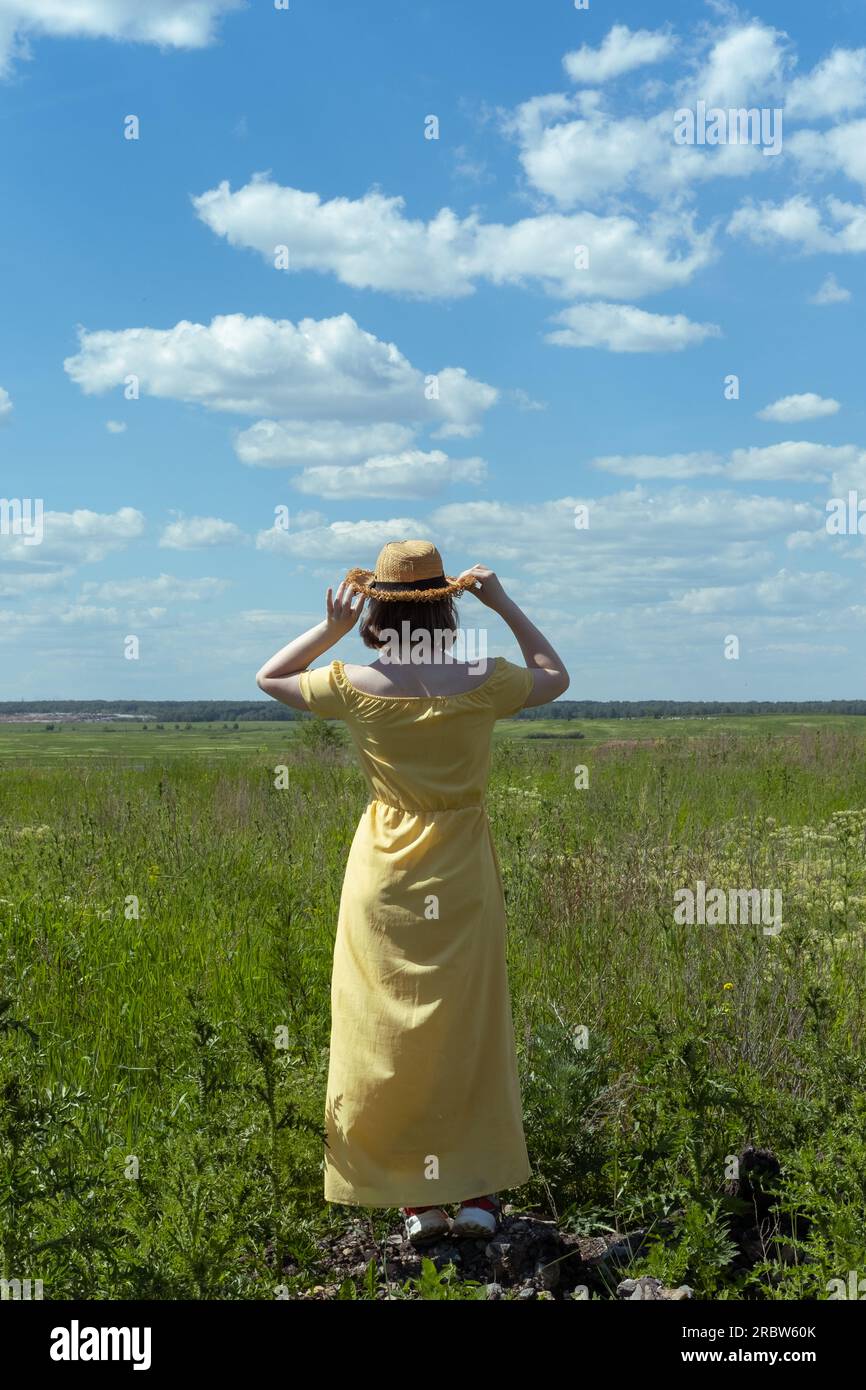 Fille asiatique dans un chapeau de paille et robe jaune parmi l'herbe moelleuse dans le champ, vue arrière. Heure d'été. Vacances au village. Temps de déplacement Banque D'Images