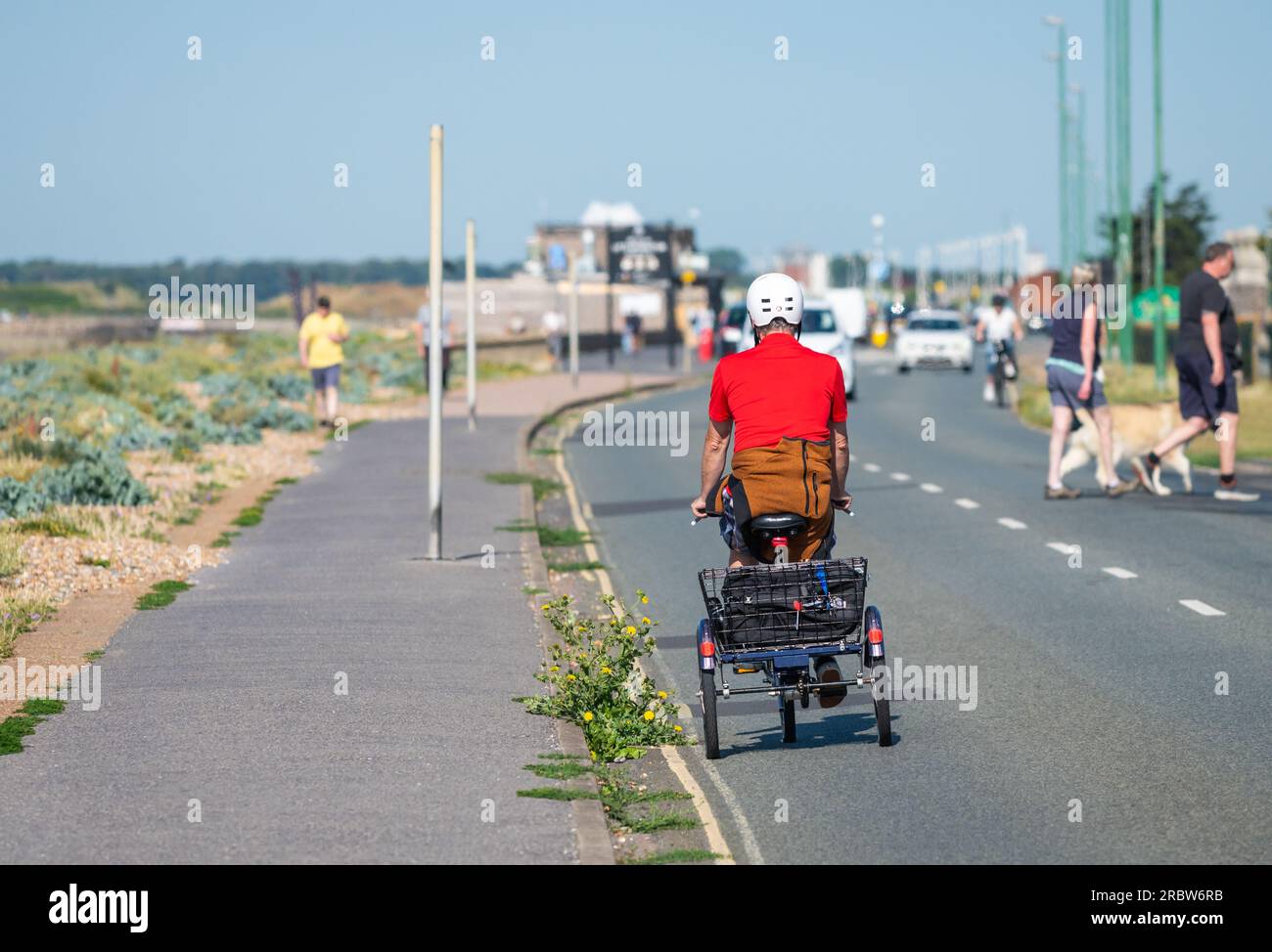 Homme de tricycle, un Pushbike avec 3 roues, avec un panier, portant un casque sur une route en bord de mer en été, Royaume-Uni. Banque D'Images