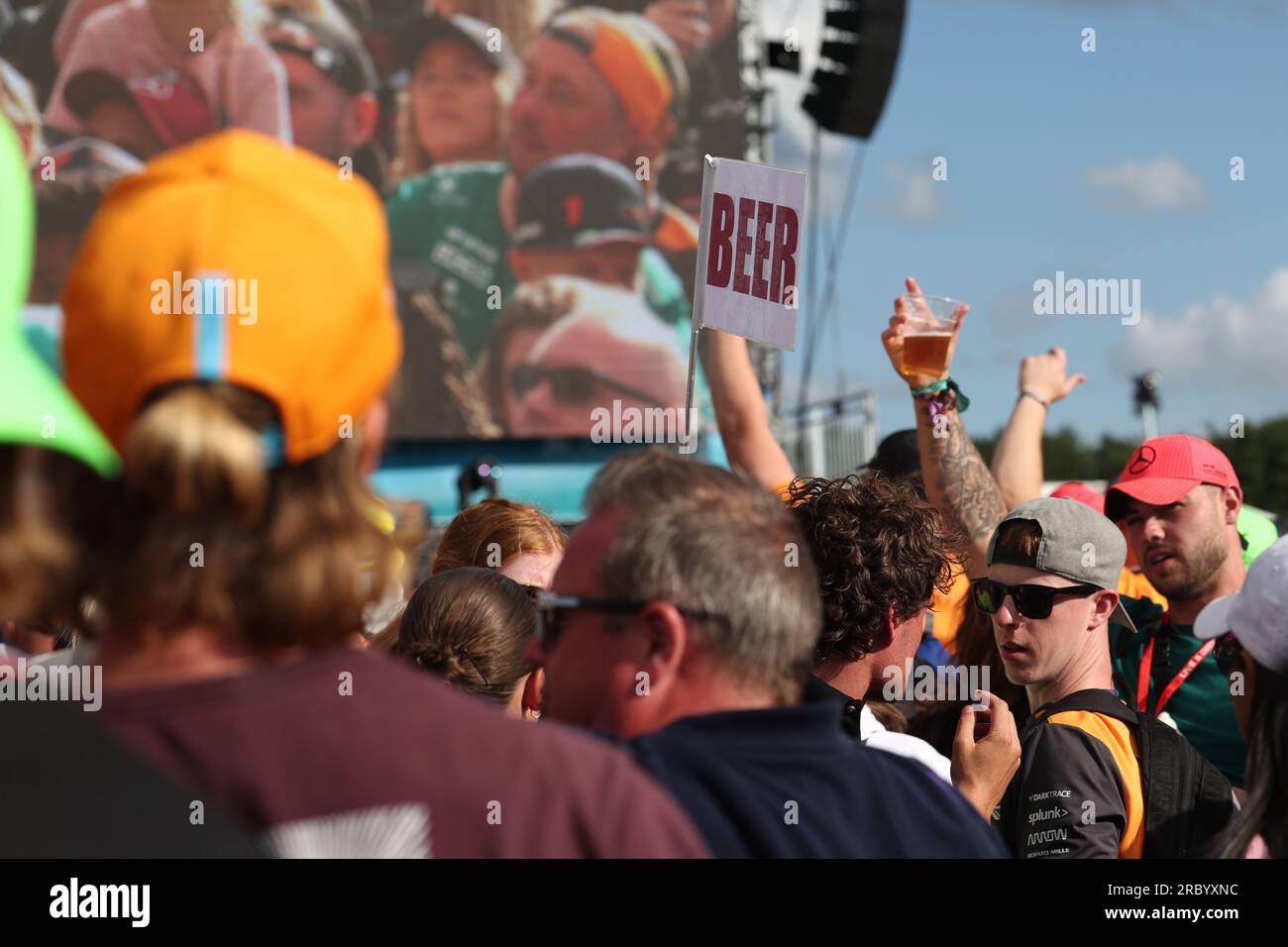 Un fan de F1 agite son verre à bière dans l'air à la musique à la fin de la journée en plein air et un vendeur de bière (le signe de bière est hissé au-dessus de la foule) tout en watc Banque D'Images
