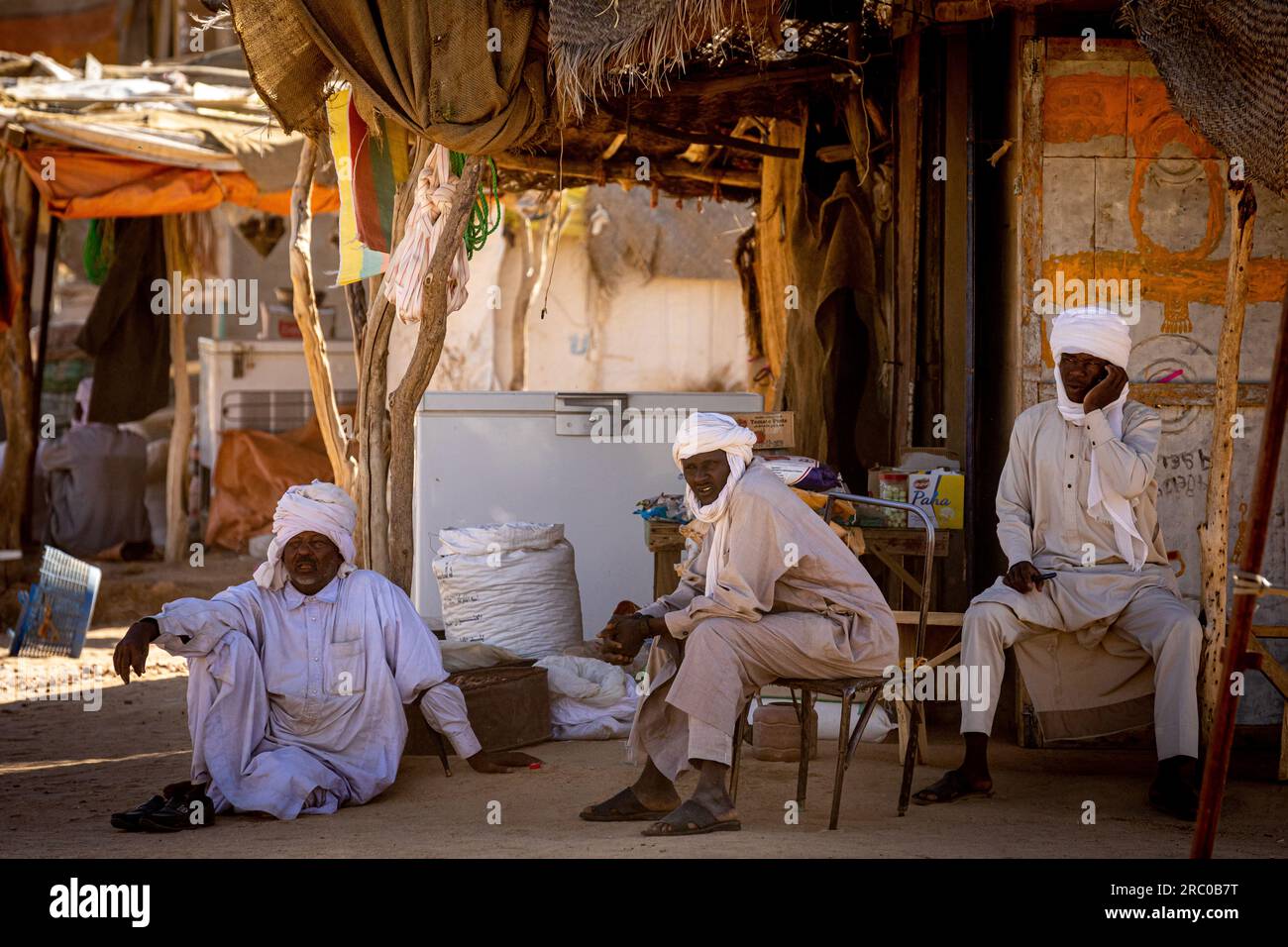 Dans le marché rural du Tchad, trois hommes vêtus de vêtements traditionnels et de turbans font du commerce sous un hangar rustique, mettant en valeur la culture locale. Banque D'Images