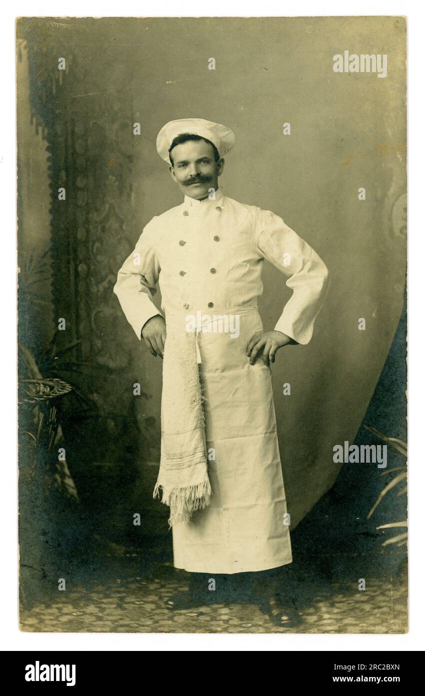 Carte postale originale du début des années 1900 d'un chef masculin joyeux, moustache, en uniforme blanc portant un chapeau de chef, serviette en tablier à la ceinture, circa 1910, Royaume-Uni Banque D'Images