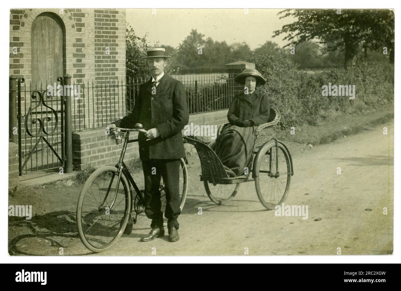 Carte postale de l'ère édouardienne du début des années 1900 d'un couple à vélo à la campagne, la femme est assise dans une remorque / chaise de bain (fauteuil roulant tôt) attachée au vélo tandis que le mari fait tout le travail. Vélo cargo tôt! Circa 1910, Royaume-Uni Banque D'Images
