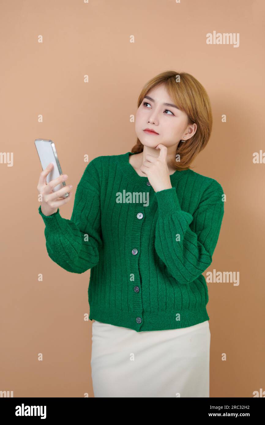 Portrait de jeune adolescente asiatique pensant sérieusement tout en tenant un téléphone portable, isolé sur fond beige Banque D'Images