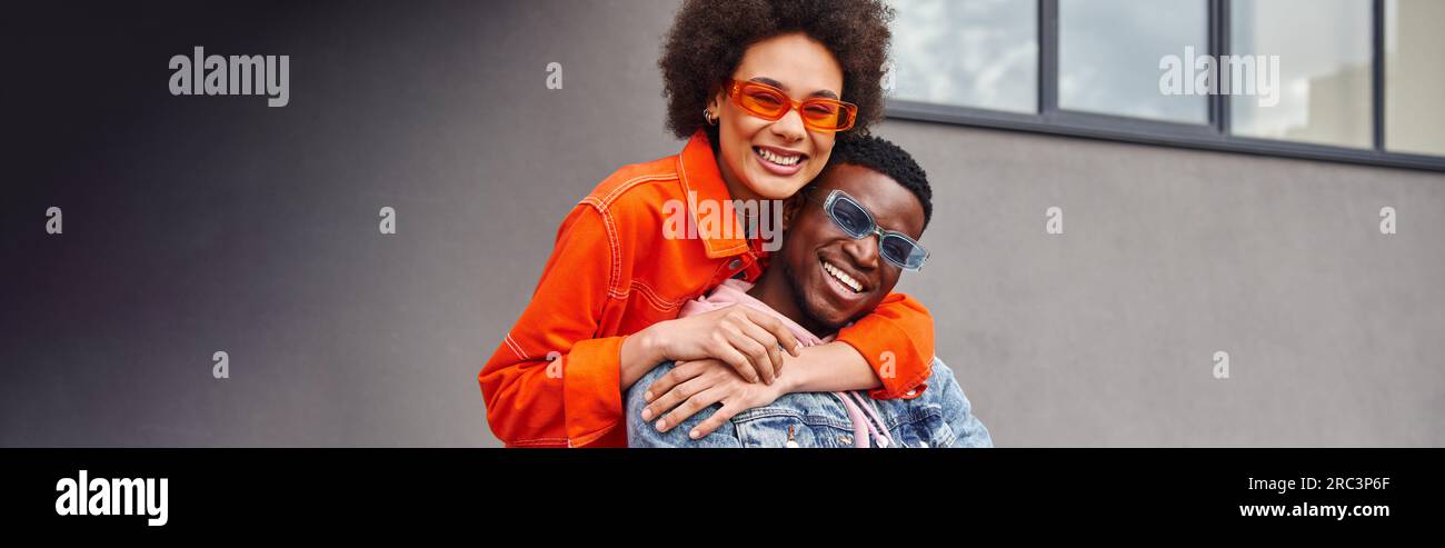 Positive jeune femme afro-américaine dans des lunettes de soleil et tenue lumineuse embrassant le meilleur ami et regardant la caméra tout en se tenant près du bâtiment sur l'urba Banque D'Images