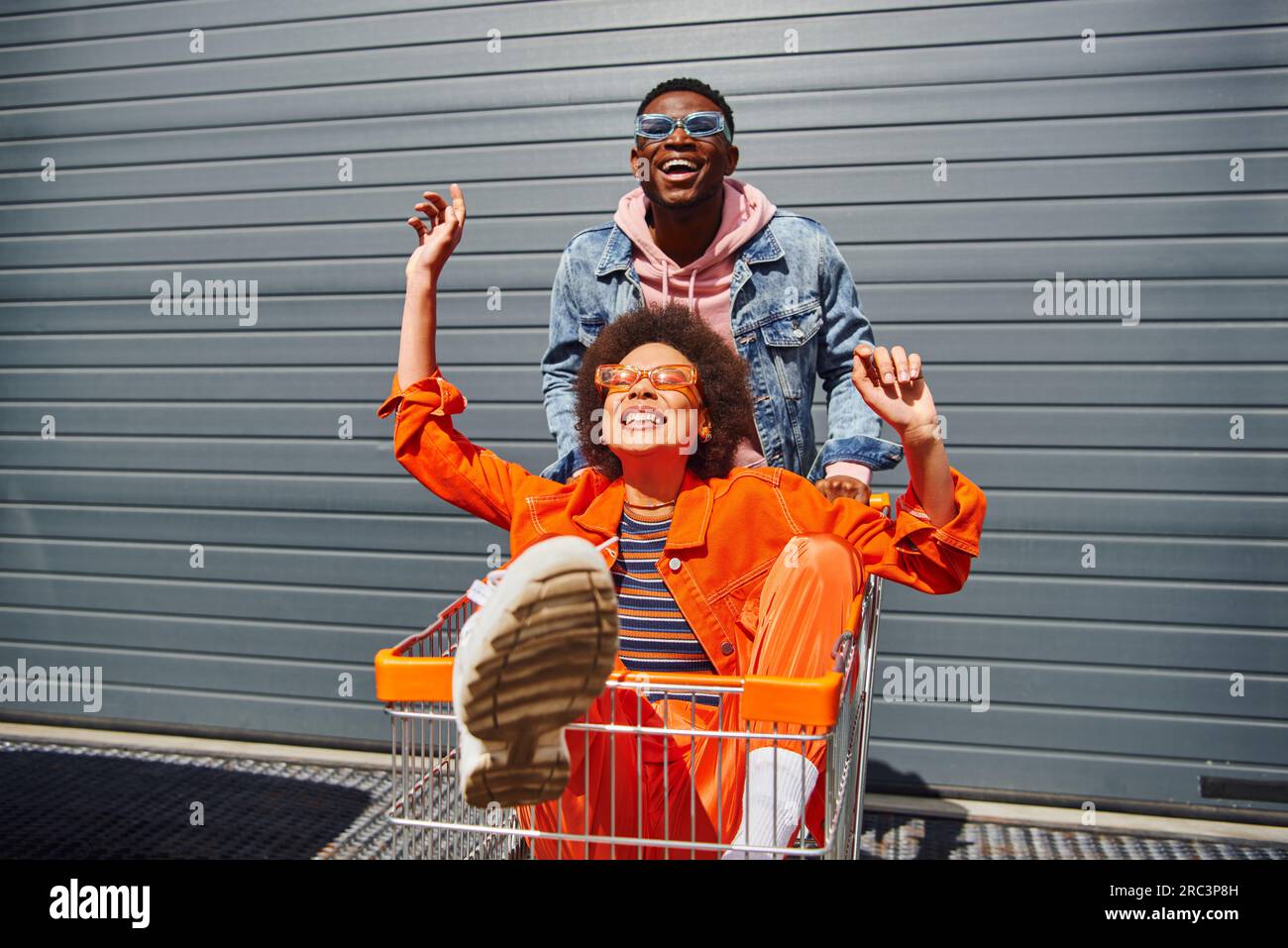 Joyeux jeune afro-américain meilleurs amis dans des lunettes de soleil et des tenues lumineuses s'amuser avec le panier et passer du temps près de la construction à backgro Banque D'Images