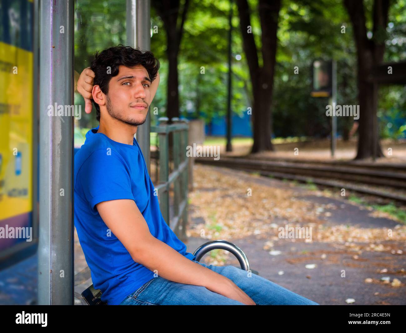 Photo de trois quarts d'un beau jeune homme en milieu urbain attendant le tram à l'arrêt de bus, portant un polo bleu Banque D'Images