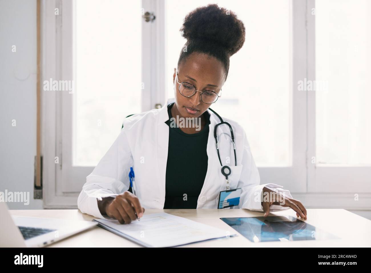 Médecin féminin ethnique avec stéthoscope assis au bureau et se concentrant tout en prenant des notes dans l'hôpital moderne Banque D'Images